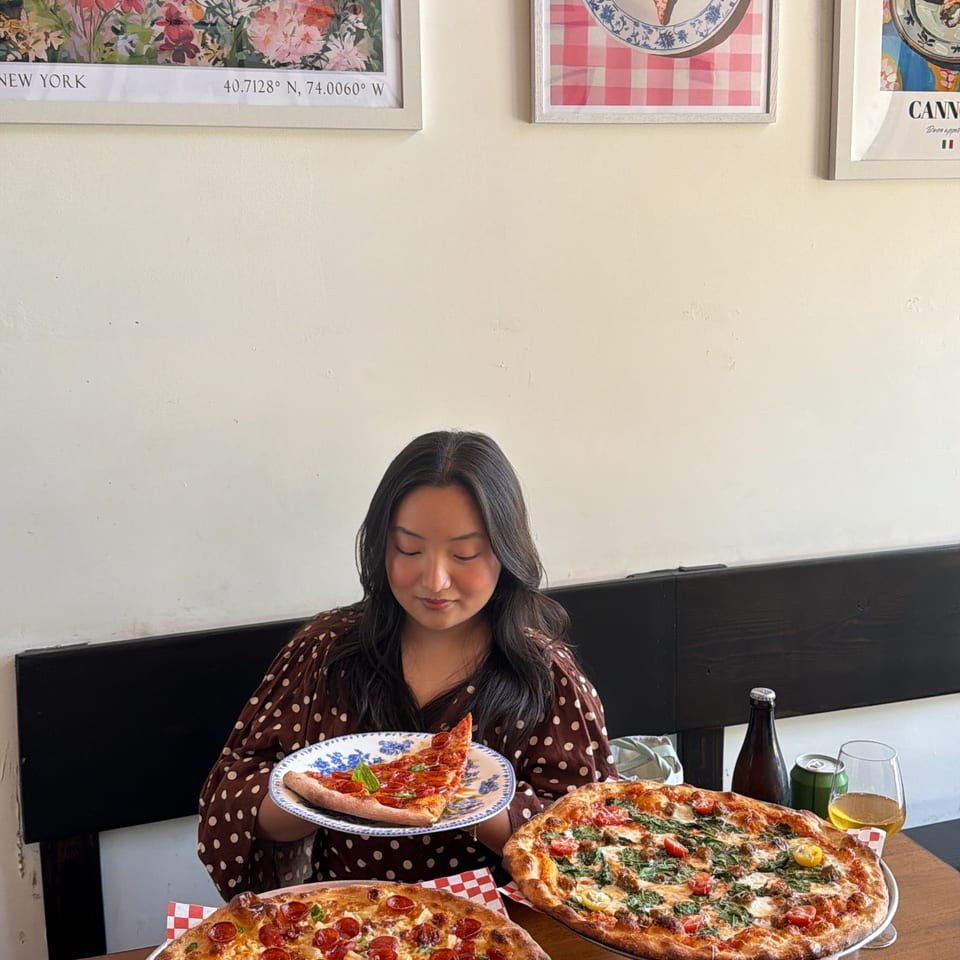 Diner holds a pizza beside three pizzas on raised stands in a cozy restaurant, beneath colorful New York and pizza artwork.