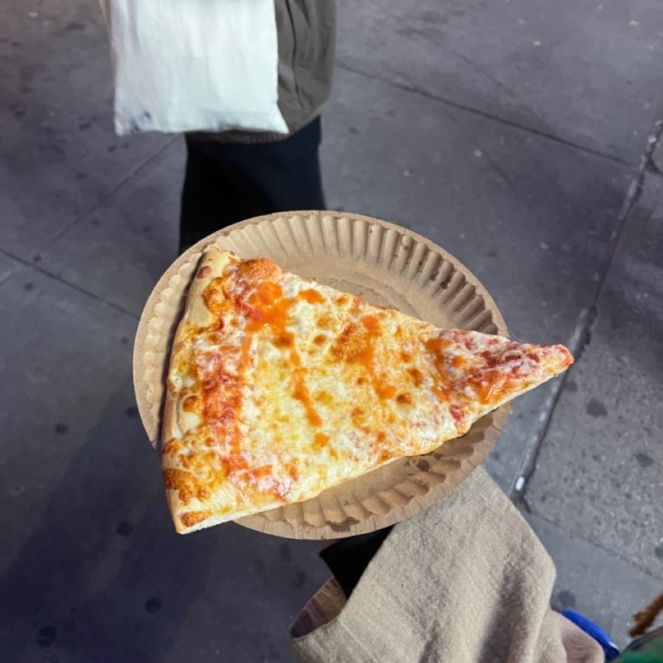 Large triangular slice of cheese pizza on a compostable paper plate, held over a city sidewalk.