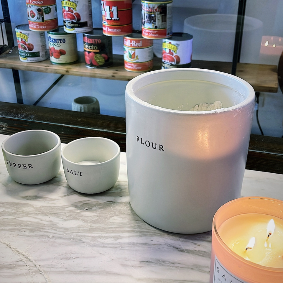 Flour, salt, and pepper containers on a marble counter with tomato cans on a shelf behind.