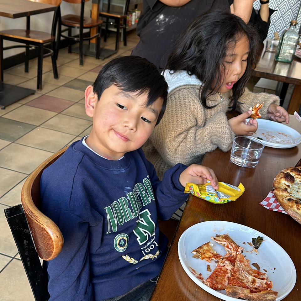 Two children eating pizza at Bloom pizzeria, with a boy in a blue sweatshirt looking at the camera.