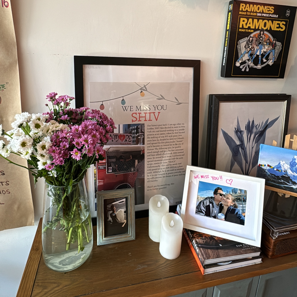 A memorial display for Shiv, featuring flowers, framed photos, candles, and a Ramones puzzle box.